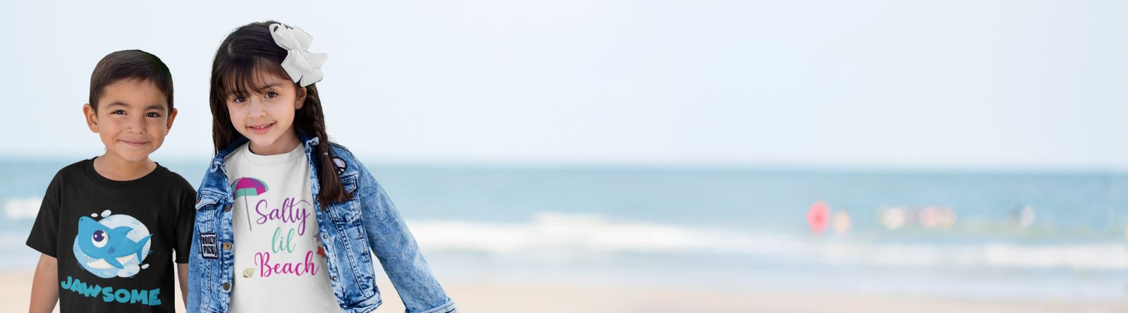 Toddler Beach Clothes features a brother and sister on the beach. She is wearing a white t-shirt saying Salty Lil Beach and his black t-shirt shows an image of a blue shark with Jawsome as the text. 