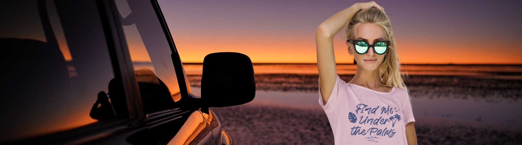 Women’s Beachwear- This image is a young woman wearing a pink t-shirt which says Meet Me Under the Palms with an image of a palm tree.  Text is in blue.  The woman is in a beach area with the shoreline visible alongside a shiny car. 
