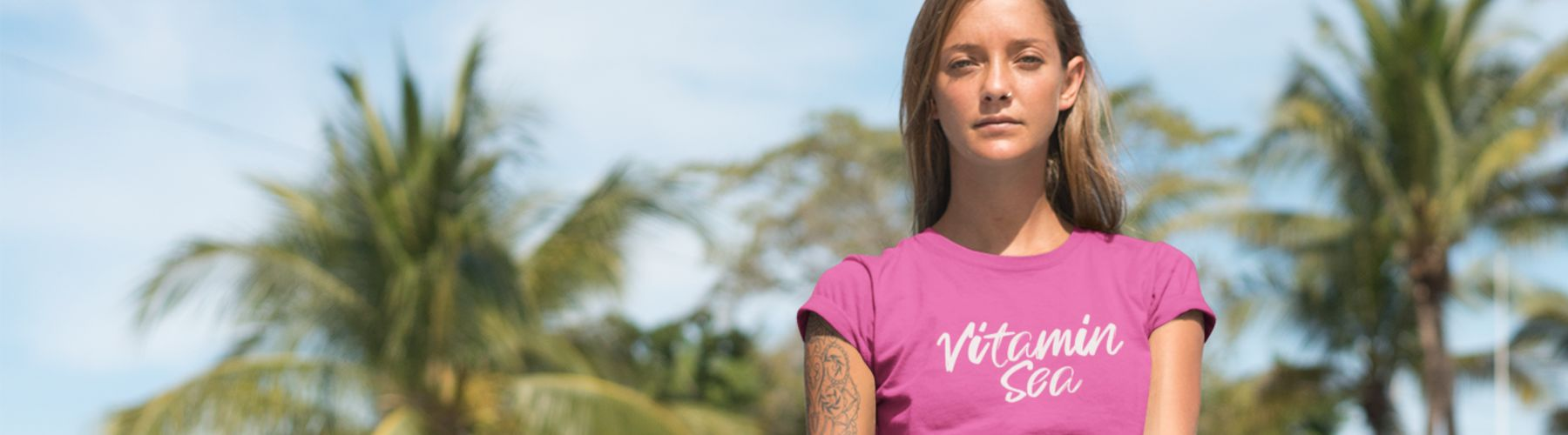 Women’s Beach T-Shirts- This image is a young woman wearing a pink t-shirt which says Vitamin Sea which is a pun on words.  The woman is in a beach area with palm trees and a blue sky. 