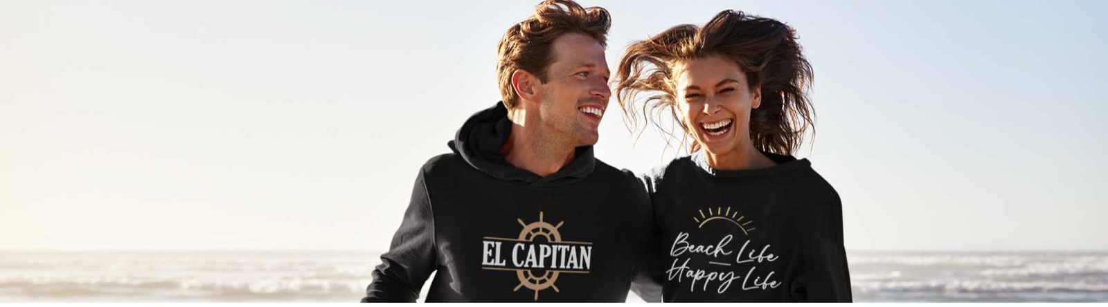 This image is a man and woman on the beach wearing a black hoodie and black sweatshirt.  The man is wearing the hoodie which says on the front El Capitan. The woman is wearing the sweatshirt which says Beach Life, Happy Life with an image of a sunset. 